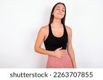 Satisfied smiling young woman wearing sportswear over white studio background , keeps hands on belly, being in good mood after eating delicious supper, demonstrates she is full. Pleasant feeling in st