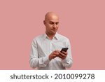 Small photo of Tired of spam. Dissatisfied bald man types message on smartphone in hands, looks angry, response to rudeness, fight with spammers. Has bristle, dressed in white shirt. Isolated over pink background