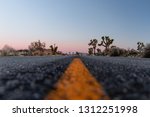 Small photo of Road less traveled, low angle viewing the yellow double lines in the desert or outdoor area. Sunset hours lighting the black paved road and mountains. Road goals to get somewhere. Joshua Tree.
