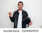 Adult Asian man wearing leather jacket holding motorcycle helmet smiling and pointing to the right side