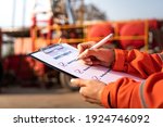Small photo of Action of safety office is writing on checklist paper during safety audit and risk verification at drilling site operation with blurred background of mount truck rig. Selective focus at hand.