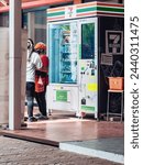 Small photo of KANCHANABURI, THAILAND - OCTOBER 4,2020 : Unidentified Asian woman wear hat, mask and apron buys drinks and snacks from 7-Eleven vending machine at PTT gas station. Convenience with vending machines.