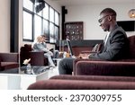 African businessman working on laptop computer while sitting on sofa in airport business lounge or hotel lobby, happy smiling male has online conference meeting during waiting flight at airport lounge