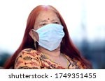 Red haired old Indian woman wearing face mask looking outside from the window. Old lady looking at outside park during Lock down. Missing pre-covid days. Missing going outside. Staying at home. 
