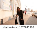 European pretty young hipster woman in a trendy black long coat in white jeans in a t-shirt posing standing in the city near the vintage white buildings. Stylish girl model enjoys the sunshine. 