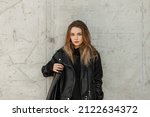 Fashionable adorable woman model with fashion black leather jacket and black hoodie with leather bag stands near a gray concrete wall