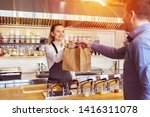 Cheerful waitress wearing apron serving customer at counter in restaurant - Small business and service concept with young woman owner offering recycled paper bag with take away food to online client