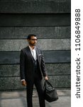 Small photo of A confident and cool Indian Asian man in a 3 piece suit is holding an exercise bag and standing against a grey wall in the day. He is wearing sunglasses and diamond earrings which makes him roguish.