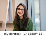 A close up head shot portrait of a preppy, young, beautiful, confident and attractive Indian Asian woman in a green sweater and spectacles in a classroom or office. She is smiling happily. 
