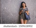Portrait of a beautiful and attractive Indian Asian young woman smiling as she checks her smartphone. She is leaning against a wall in a city in the day and is smiling as she uses her device.