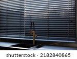 Small photo of Wood blinds black color closeup on the window. Wooden slats 50mm wide. Venetian bamboo blinds in the kitchen. Black tapes. Sink with copper faucet near the window. Stone countertop.