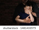 Adult man covering scared little boy's mouth near brick wall, space for text. Child in danger