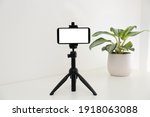 Smartphone with blank screen fixed to tripod on white table indoors. Mockup for design