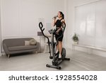 Happy young woman drinking water during training on elliptical machine at home