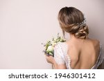 Young bride with elegant hairstyle holding wedding bouquet on beige background, back view. Space for text