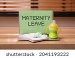 Baby socks, feeding bottle, notebook and laptop with text Maternity Leave on wooden table indoors