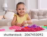 Small photo of Cute little girl playing with bright kinetic sand at table in room