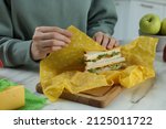 Small photo of Woman packing tasty sandwich into beeswax food wrap at white marble table indoors, closeup