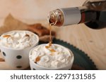 Pouring delicious caramel syrup into cup with coffee and whipped cream at wooden table, closeup