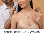 Small photo of Man putting elegant necklace on beautiful woman against beige background, closeup