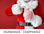 Santa Claus putting coin into piggy bank on color background, closeup