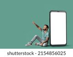 Cool tattooed young man sitting near big smartphone and pointing at something on green background