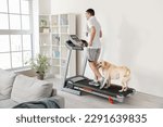 Small photo of Young man with cute Labrador dog training on treadmill at home
