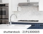 Small photo of A luxury kitchen sink detail shot with marble countertops, herringbone tile backsplash, and white and blue cabinets.
