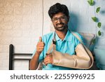 Small photo of happy smiling young man with broken hand at bedroom showing thumbs up gesture by looming at camera - concept of recovered, successful medical treatment and rehabilitation
