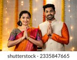 Happy young indian couple in traditional ethnic clothing saying namaste or welcome gesture by looking camera during festival celebration at home - concept of greeting, wishes and togetherness.
