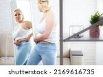 Small photo of Displeased middle-aged woman looking at herself in the mirror and pinching stomach fat in domestic interior. Sad plus size woman looking her reflection in the mirror with sorrow at home.