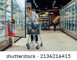 Small photo of Young beautiful mother holding pushing shopping cart with her child in supermarket. Girl is choosing daily milk product picking up from shelf with her mother beside. Shopping for healthy.