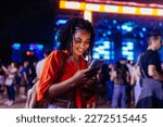 A young African American girl is with a smartphone at a music festival, texting with her friends.