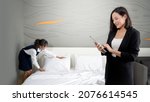 Small photo of Asian businesswoman in black suit standing with tablet computer in her hand. The hotel manager verify the tidiness of the room for hotel guests while the maid making the bed in the background.