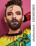 Small photo of Male makeup look. Portrait of a young man in colorful shirt, wearing beard, quiff, purple lipstick, eyeliner, pink eyeshadow. The guy looking at the camera over pink background, one hand on his nape.