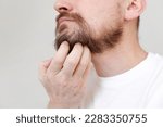 Small photo of Cropped shot mans hand is unrestrained scratching unshaven chin. Overgrown unkempt male in white t shirt with thick beard and mustache face. Beauty salon concept, barbershop.