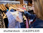 Small photo of Young Woman Buying Used Sustainable Clothes From Second Hand Charity Shop Looking At Price Tag