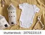 Small photo of Styled stock photo of clear white baby onesie with scandinavian stitched pillow in the shape of a house and wooden toys on yellow napkin for creating mockup for presentation kids sublimation design