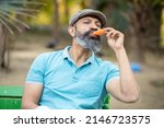 Indian senior man eating licking ice lolly or ice cream in a park outdoor, mature bead male enjoy retirement life. summer holidays. selective focus