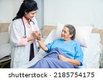 Smiling Indian caring doctor supporting holding hand of olde senior female patient lying on bed at clinic or hospital. Elderly people health care concept.