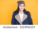 Portrait of happy young indian girl wearing winter cloths smiling isolated over orange yellow background. She is wearing woolen hat and blue jacket or over coat.
