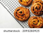Just baked pain aux raisins on cooking wire rack. Buns are also called escargot or pain russe, is a spiral pastry with custard cream and raisin. Directly above, white table surface.