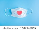 Small photo of Disposable medical mask on blue background with red heart on it against bacteria and viruses. Stop the spread of coronavirus, self-defense. Copy space