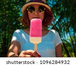 Beautiful Woman Short Hair Wearing White Shirt, Hat and Round Sunglasses with Happy Smiling Holding White and Pink Frozen Popsicle Ice Pop on Summer Time with Sunshine on Tree and Blue Sky Background