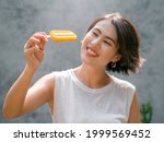 Yellow popsicle in beautiful happy Asian woman’s hand wearing casual white sleeveless shirt, outdoors. Attractive woman showing popsicles. Smiling female enjoying ice lolly in summer.