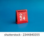 Small photo of Happy new year 2024 banner background. 2024 numbers year with target dart icon on red small desk calendar cover standing on blue background, minimal style. Business goals plan and success concepts.