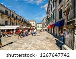 Small photo of Kotor, Montenegro - A young woman selling souvenirs takes a cigarette break in the shade in the Piazza of the Arms in the ancient city of Kotor, Montenegro as tourists walk by the cafes and shops