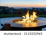 A woman reclines on a hillside patio terrace with her feet up on the edge of a flaming fire pit as the sun sets and the city lights go on in the distant view.