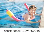 Active senior (elderly) woman (over age of 50) in sport goggles, swimsuit doing aqua fitness with swim noodles in swimming pool. Mature female smiling with happy face in summer day. Healthy lifestyle.