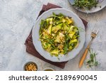A healthy salad of raw zucchini, cheese, a mixture of microgreens and roasted almond slices with mustard dressing on a gray ceramic plate on a light concrete background. Salad recipes. Healthy food